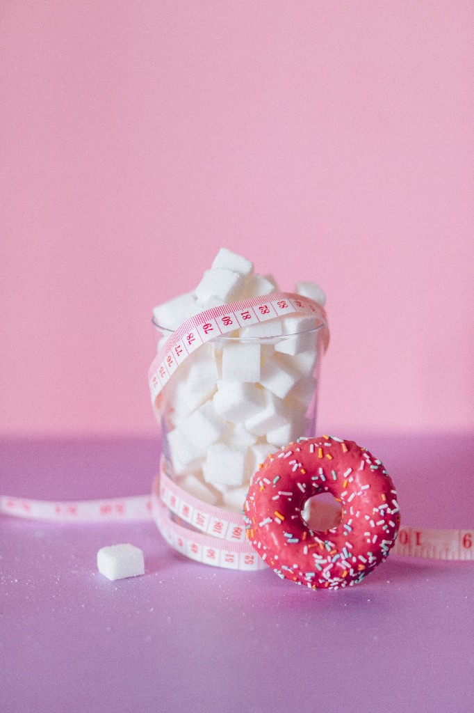 Glas mit Zuckerwürfeln, Maßband und ein Donut auf rosa Grund